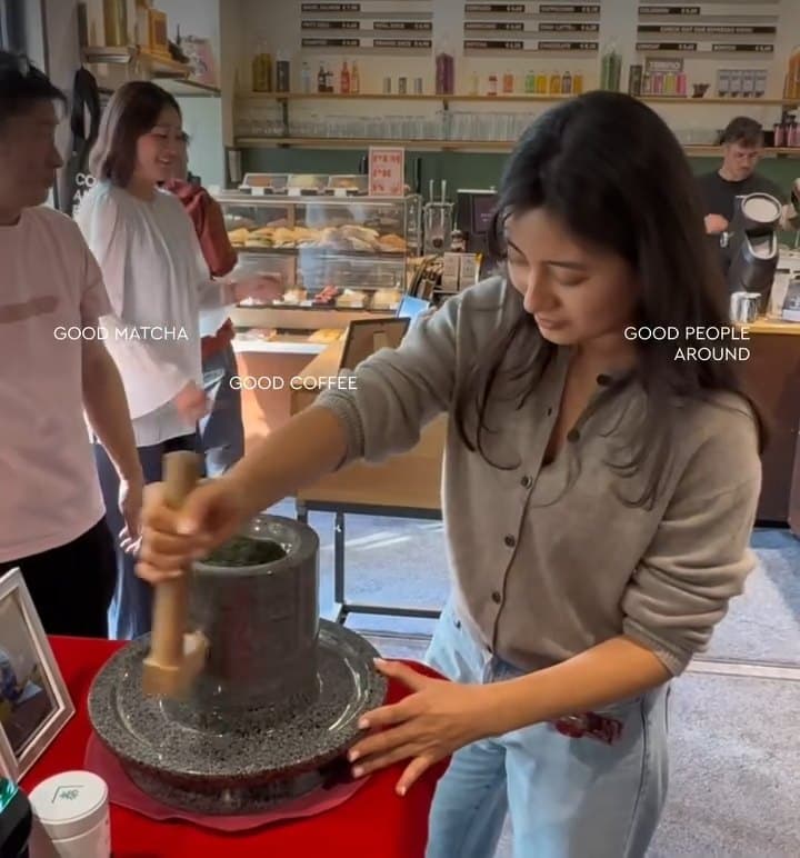 A woman turns the wooden handle of a stone mill on a red cloth
